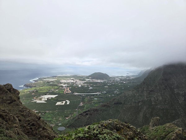 Vue en hauteur de Buenavista del Norte avec des champs verdoyants, des serres et le littoral sous les nuages et la brume.
