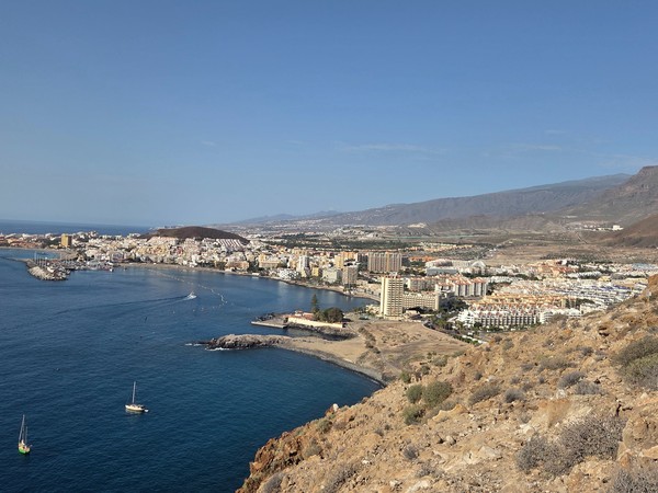 Vue panoramique de Los Cristianos et du littoral depuis un point élevé, avec la mer calme, les hôtels et les montagnes en arrière-plan.