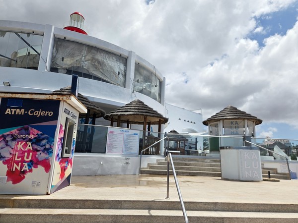 Entrée du Kaluna Beach Club avec un distributeur automatique, des escaliers et des terrasses blanches sous un ciel nuageux.