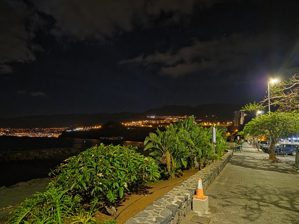 Promenade nocturne au bord de la mer à Radazul avec lampadaires, plantes et lumières de la côte au loin.