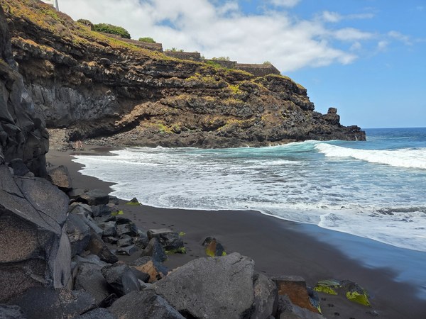 Petite crique de la plage d’Ancón à Tenerife, avec sable noir, rochers volcaniques, falaise abrupte et vagues blanches.