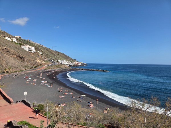 Vue de la plage de La Nea à Tenerife, avec sable noir, eau calme, digue de protection et collines en arrière-plan.