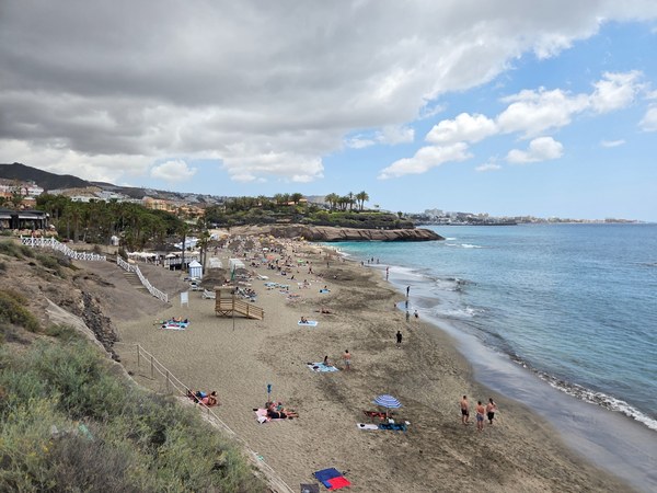 Vue large de Playa El Duque à Tenerife, avec plage animée, eau calme et promenade côtière sous un ciel nuageux.