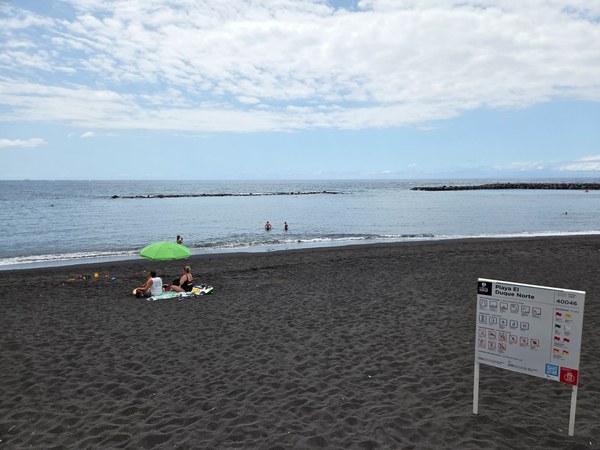 El Duque Nord Secteur nord de Playa El Duque à Tenerife, avec sable noir, mer tranquille et panneau d’information au bord de la plage.