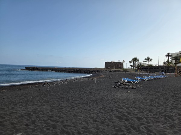 Playa Jardín à Tenerife, avec sable noir, vagues blanches, falaises côtières et quelques personnes sur la plage.