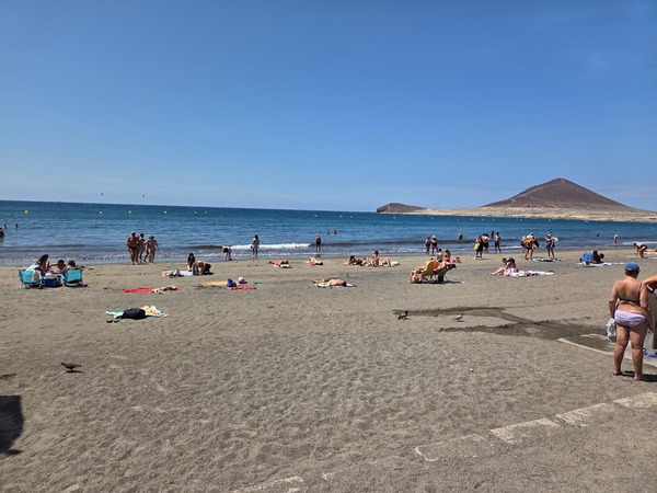 Plage de sable sombre à El Médano avec des baigneurs, la mer calme et la Montaña Roja en arrière-plan.