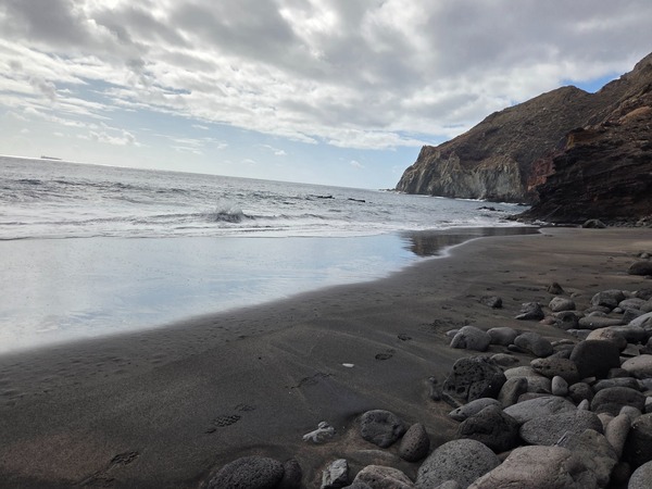 Rivage de sable noir avec galets, falaise volcanique et mer calme sous un ciel nuageux.