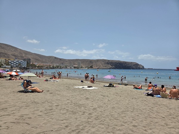 Plage urbaine de Los Cristianos avec nombreux baigneurs, eau turquoise et collines arides au fond.