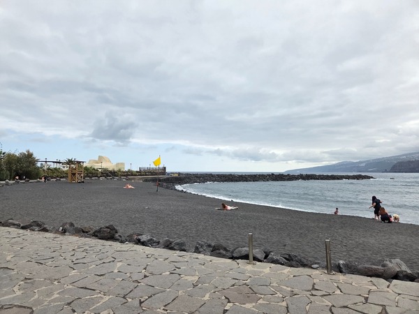 Plage de sable noir près de Puerto de la Cruz avec drapeau jaune, mer tranquille et ciel couvert.