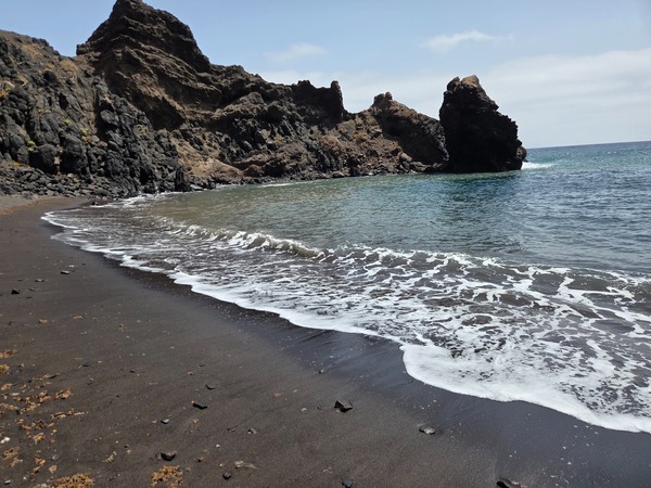 Petite crique de sable noir à Roque Bermejo avec rochers volcaniques abrupts et eau claire.